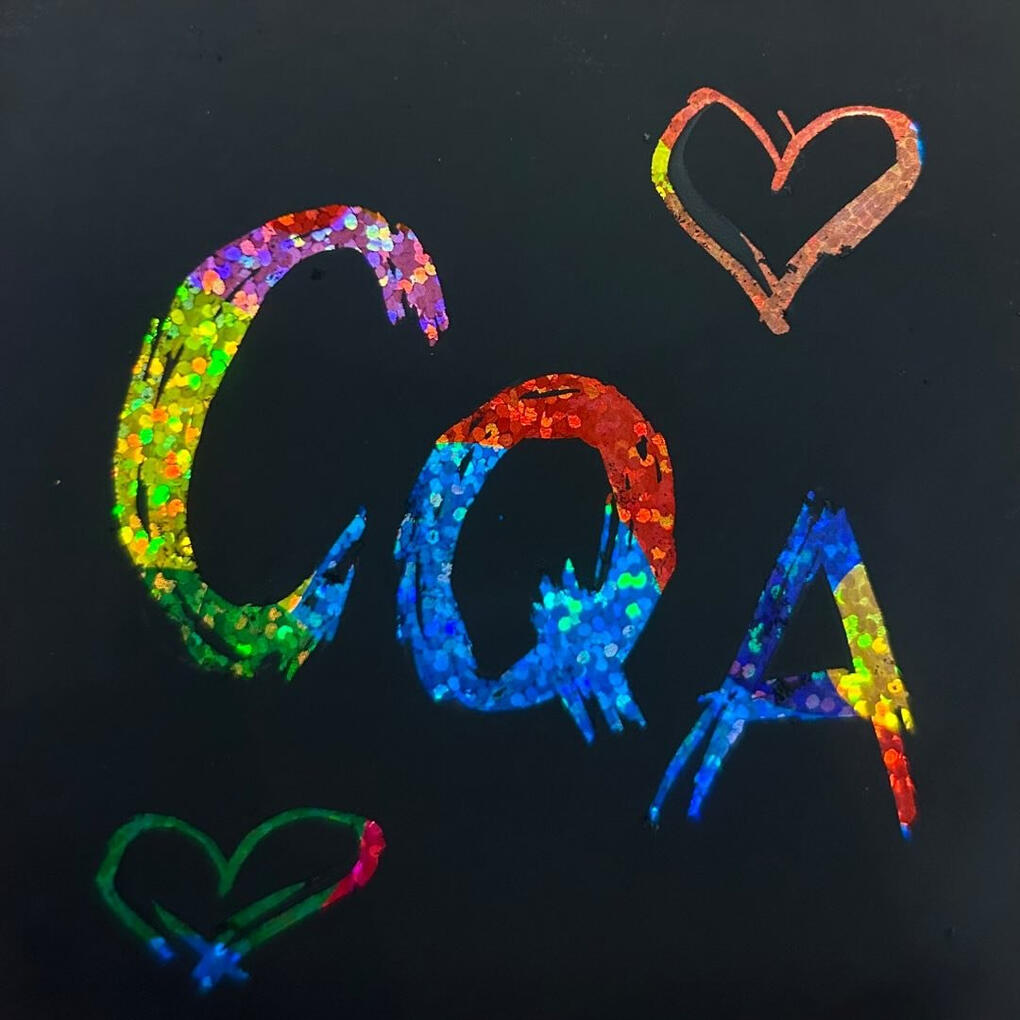 Glittery rainbow letters scratched into a black surface reading C Q A and surrounded by two hearts.